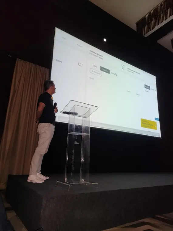 Fabricio Costa no palco de um auditório, apresentando um sistema projetado em tela grande